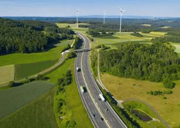 Aerial view of a highway cutting through a green landscape with wind turbines, showcasing sustainable logistics. - Supply Chain News