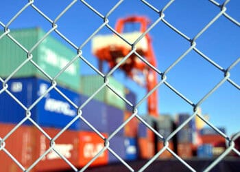A chain link fence with containers in the background, symbolizing the pervasive challenges associated with illegal trade and the need for enhanced intelligence sharing through technological advancements.