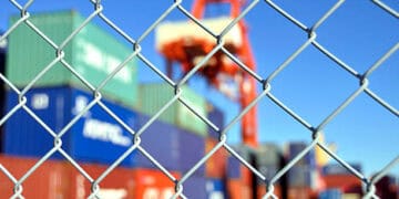 A chain link fence with containers in the background, symbolizing the pervasive challenges associated with illegal trade and the need for enhanced intelligence sharing through technological advancements.