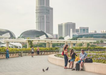 A group of people sitting on a bench in front of an expanding skyscraper in Singapore, discussing anti-money laundering measures and the ownership of luxury assets.