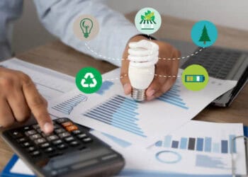 Person reviewing sustainability data, holding energy-efficient lightbulb. Corporate Social Responsibility. - Supply Chain News