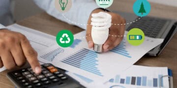 Person reviewing sustainability data, holding energy-efficient lightbulb. Corporate Social Responsibility. - Supply Chain News