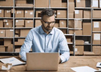 Man using laptop in warehouse, improving supply chain efficiency. - Supply Chain News