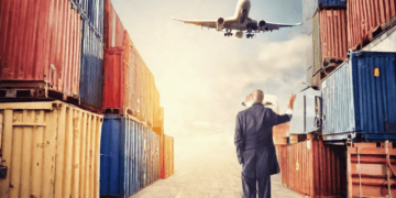 Businessman watching airplane landing near stacked cargo containers. Global supply chain data. - Supply Chain News