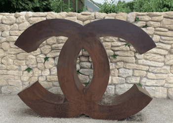 Large rusted metal Chanel logo in front of a stone wall. - Supply Chain News