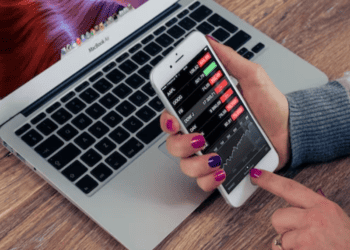 Woman using smartphone to track stock market data, laptop nearby. - Supply Chain News