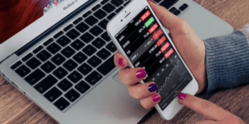 Woman using smartphone to track stock market data, laptop nearby. - Supply Chain News