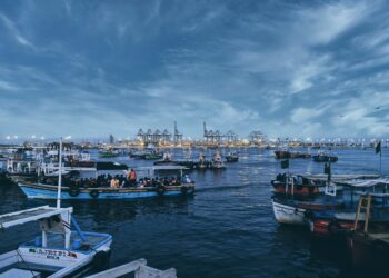 Fishing boats at dusk near a busy port, showcasing the contrast between local life and global trade. - Supply Chain News
