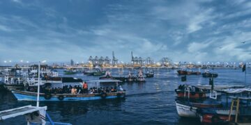 Fishing boats at dusk near a busy port, showcasing the contrast between local life and global trade. - Supply Chain News