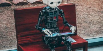 AI robot sitting on a bench, using a laptop. AI100 progress report. - Supply Chain News