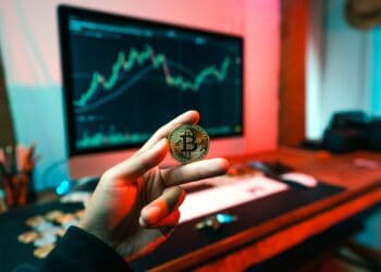 Person holding a Bitcoin cryptocurrency coin against a background of a computer screen showing a stock chart. - Supply Chain News
