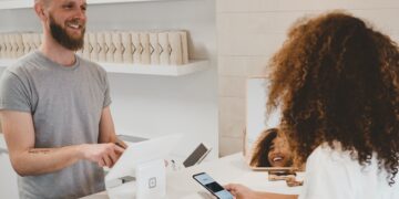 Smiling shopkeeper helps customer with contactless payment using a smartphone. - Supply Chain News