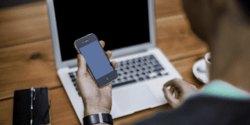 Person working on laptop, holding smartphone displaying blank screen. Digital passport concept. - Supply Chain News