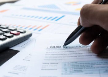 Close-up of a hand filling out a 1040 tax form with a pen. - Supply Chain News