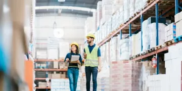 Two warehouse workers in hard hats review paperwork, showcasing efficient freight forwarding operations for 2023. - Supply Chain News