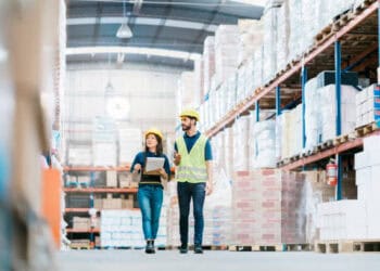 Two warehouse workers in hard hats walking through a warehouse to prepare for the holiday season.