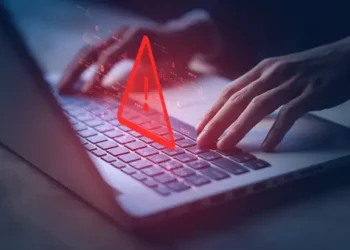 Hands typing on laptop with a red warning triangle overlay. Cybercrime treaty impact. - Supply Chain News