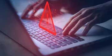 Hands typing on laptop with a red warning triangle overlay. Cybercrime treaty impact. - Supply Chain News