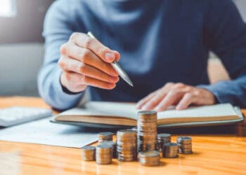 Person reviewing finances, stacks of coins on desk near open notebook and pen. - Supply Chain News