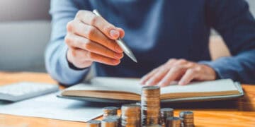 Person reviewing finances, stacks of coins on desk near open notebook and pen. - Supply Chain News