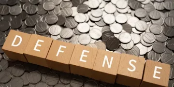 Wooden blocks spelling "DEFENSE" on a pile of coins. Japan's defense budget. - Supply Chain News