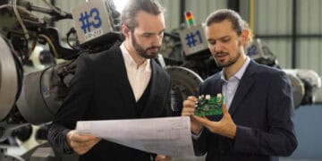 Two engineers review a circuit board and blueprints, assessing risk in a factory setting. #3, #1 - Supply Chain News