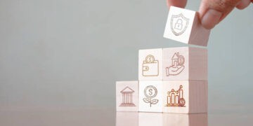 A hand is holding wooden blocks with business icons on them, showcasing the impact of US-EU trade policies in the Biden era.