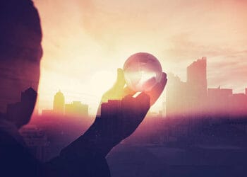 Silhouette of a hand holding a globe, superimposed over a city skyline at sunset. Global economic growth potential. - Supply Chain News