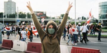 Woman at protest raising hands in peace sign, wearing a mask. - Supply Chain News