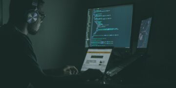 Person wearing headphones works on laptop and monitors displaying code, boosting cybersecurity skills for Moldovan SMEs. - Supply Chain News