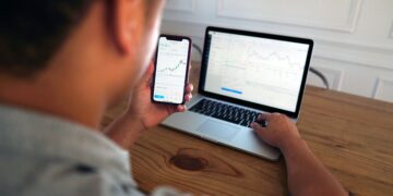 Person reviewing financial data on a laptop and smartphone. Stock market charts are visible. - Supply Chain News