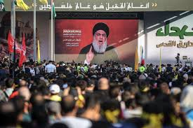 Large crowd at a Hezbollah rally, watching a large screen showing Hassan Nasrallah addressing cross-border fighting with Israel. - Supply Chain News