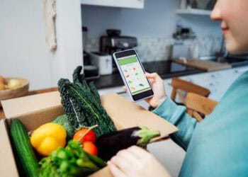 Person using smartphone to order groceries, then placing fresh produce into a box for delivery. - Supply Chain News