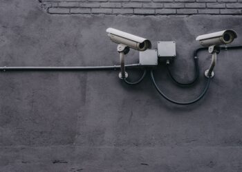 Two security cameras mounted on a wall, connected by wires and junction boxes. AI's role in cybersecurity. - Supply Chain News