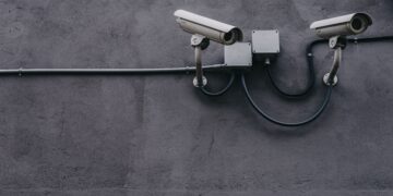 Two security cameras mounted on a wall, connected by wires and junction boxes. AI's role in cybersecurity. - Supply Chain News