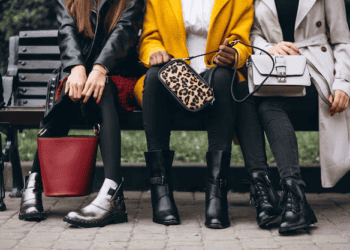 Three women wearing boots and stylish handbags sit on a park bench. Luxury fashion accessories. - Supply Chain News
