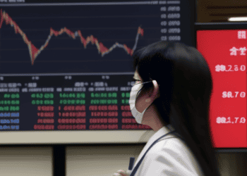 A woman wearing a face mask in front of a screen showing Asia Markets.