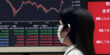 A woman wearing a face mask in front of a screen showing Asia Markets.