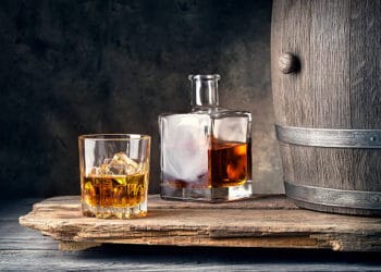 An American whiskey barrel sits on a wooden table, as a glass of whiskey is poured delicately beside it.