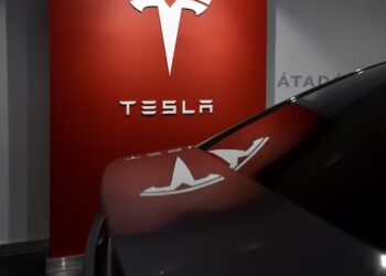Tesla logo on a red wall with a Tesla car's rear end reflecting the logo. - Supply Chain News
