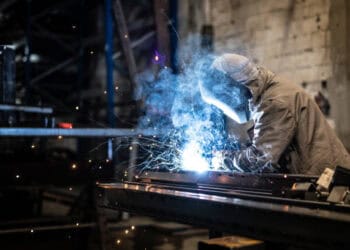 A welder is welding metal in a factory, working alongside steelworkers.