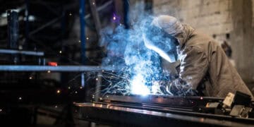 A welder is welding metal in a factory, working alongside steelworkers.