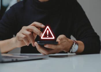 A man using a smartphone with a red warning sign on it, alerting him about the potential impact of online fraud.