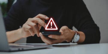 A man using a smartphone with a red warning sign on it, alerting him about the potential impact of online fraud.