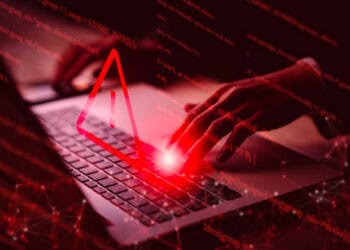 Hands typing on laptop, red warning triangle overlay. Medibank cyber attack. - Supply Chain News