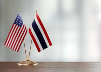 Two American and Thai flags on a wooden table representing the diplomatic relations between the two nations.
