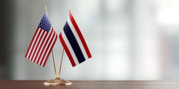 Two American and Thai flags on a wooden table representing the diplomatic relations between the two nations.