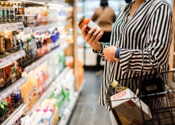 Woman examining a jar of pasta sauce in a grocery store aisle, impacted by ending tariff exemptions. - Supply Chain News