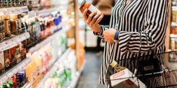 Woman examining a jar of pasta sauce in a grocery store aisle, impacted by ending tariff exemptions. - Supply Chain News