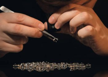 Close-up of gemologist inspecting a diamond with tweezers and loupe, surrounded by loose diamonds. Impact on global supply chain. - Supply Chain News